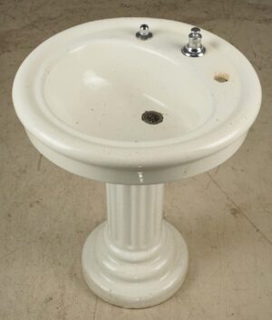White ceramic pedestal sink with a fluted column base, shown from above with two faucet openings, a corroded drain, and visible surface wear and chips.