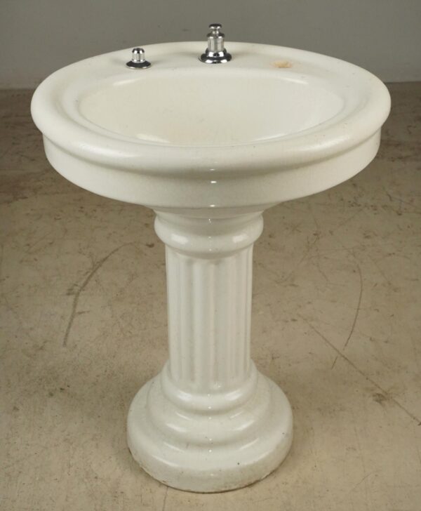 White ceramic pedestal sink with a fluted column base and two faucet openings, shown with remaining chrome hardware on top.
