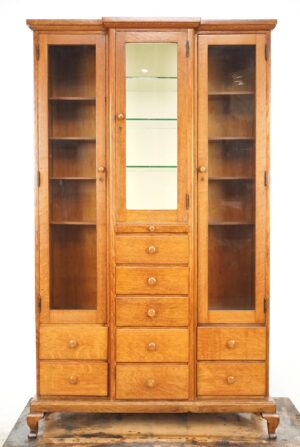 Antique oak medical storage cabinet with glass doors, central mirrored door, nine drawers, and a base with modest Queen Anne style legs.