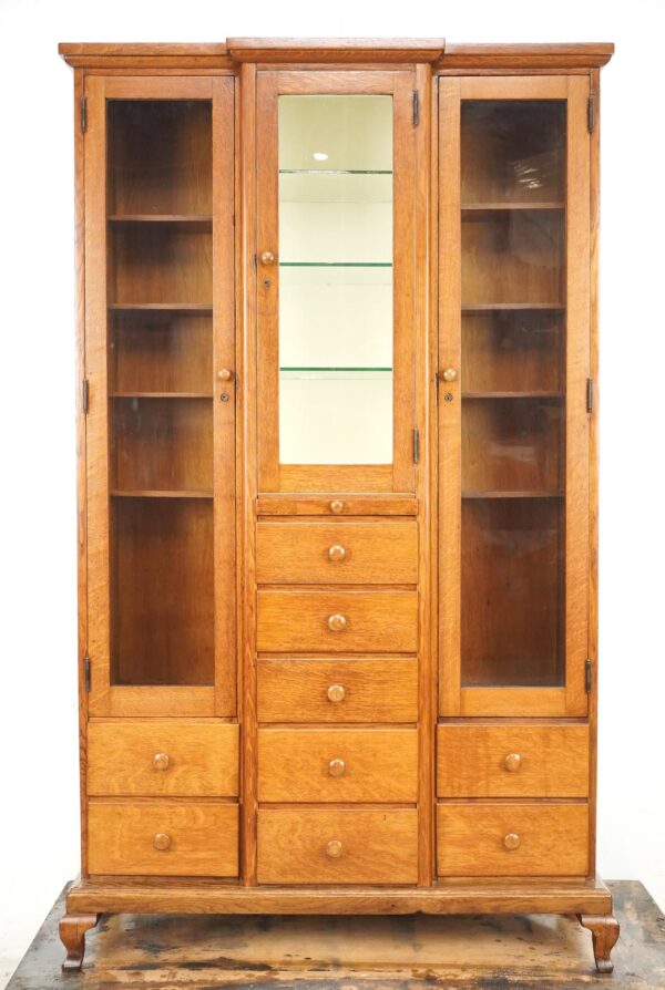 Antique oak medical storage cabinet with glass doors, central mirrored door, nine drawers, and a base with modest Queen Anne style legs.