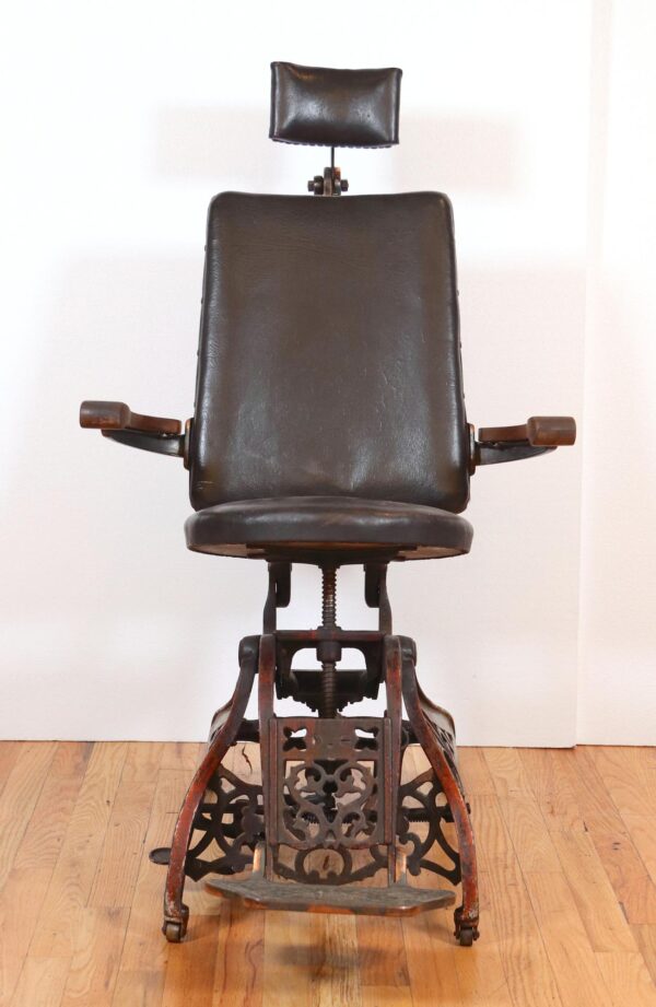 Antique examination chair with a black vinyl seat and backrest, wood armrests, and a cast iron base with foot rest shown from the front.