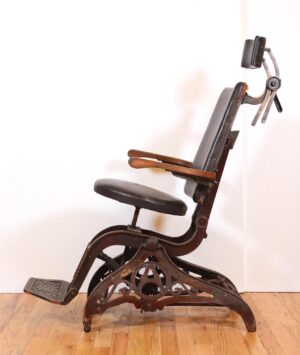 Antique examination chair with vinyl seat and back, wood armrests, cast iron base, and footrest shown in profile.