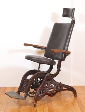Antique examination chair with a cast iron base, vinyl seat and back, wood armrests, and a foot rest.