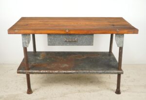 Industrial workbench with a medium wood tabletop, galvanized steel drawer and lower shelf, shown front view with iron legs and visible wear.