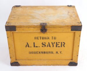 Antique yellow wooden bakery box with iron corner reinforcements and a rusted front latch, shown closed with the stenciled label Return to A. L. Sayer Ogdensburg, N. Y. visible on the side.