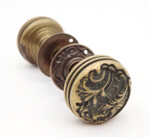 Ornate brass and bronze door knob set with rosettes and spindle, shown in close-up with one knob face featuring an asymmetrical Rococo floral pattern.