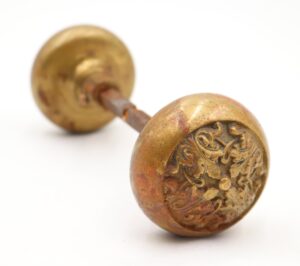 Pair of brass door knobs with a spindle, showing one knob in close-up and the other blurred behind it, with ornate German Renaissance-style detailing and visible surface wear.