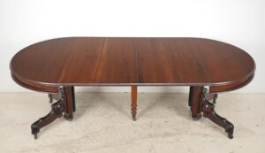 Victorian-style expandable dining table with a dark wood tone, shown extended with rounded ends, a turned pedestal center, and carved splay legs on casters.