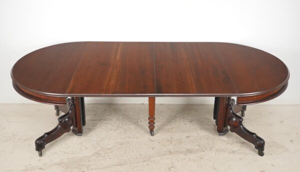 Victorian-style expandable dining table with a dark wood tone, shown extended with rounded ends, a turned pedestal center, and carved splay legs on casters.