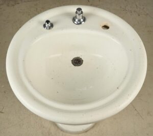 Vintage Art Deco white ceramic pedestal sink with two faucet openings, a drain, and surface wear.