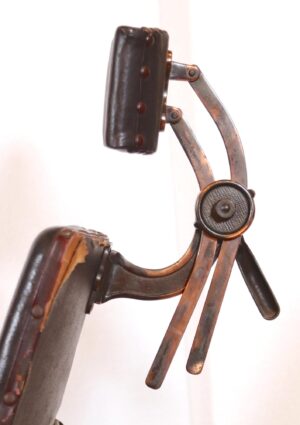 Close-up of the chair’s arm and adjustment hardware, showing the black vinyl pad, medium wood arm, and copper-toned cast iron joints.