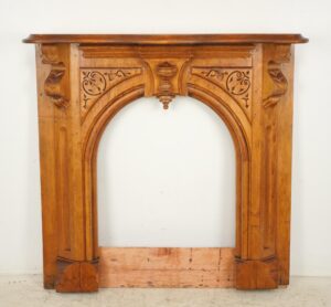 Late Victorian wooden mantel in medium wood tone, shown front on with an arched opening, carved scrollwork and geometric details, and visible surface wear along the base.